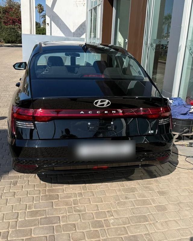 Hyundai Accent noire de l’agence, vue arrière, devant un concessionnaire Hyundai ; plaque provisoire floutée